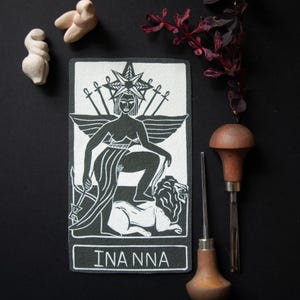 May include: A black and white tarot card with the name "INANNA" at the bottom. The card depicts a woman with a crown, wings, and swords, seated on a lion. Two small sculptures and carving tools are also present.
