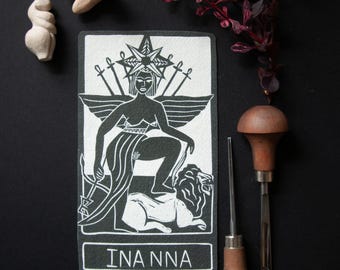 Pode incluir: Uma carta de tarô em preto e branco com o nome "INANNA" na parte inferior. A carta retrata uma mulher com coroa, asas e espadas, sentada em um leão. Duas pequenas esculturas e ferramentas de entalhe também estão presentes.