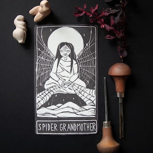 May include: A black and white illustration of a seated figure with long hair, surrounded by a spiderweb design, and a full moon in the background. The text "SPIDER GRANDMOTHER" is at the bottom. The image is accompanied by two small sculptures and two wood carving tools.