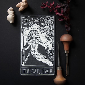 May include: A black and white tarot card featuring an illustration of a woman with long hair, holding a staff, and the text "THE CAILLEACH". The card is surrounded by other objects, including a carving and carving tools.