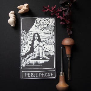 May include: A black and white Persephone art print depicting a seated woman beneath a tree. The print is displayed with two small, off-white sculptures, a sprig of red leaves, and two wood-handled carving tools. The word "PERSEPHONE" is printed at the bottom of the art print.