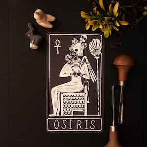 May include: Black and white art print of Osiris, an Egyptian deity, seated on a throne. The print features intricate details and the word "OSIRIS" at the bottom. The image includes a small sculpture and carving tools.
