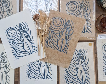 May include: A collection of blue and white linocut prints featuring a stylized female figure with long hair, standing with arms raised. The prints are on brown paper and white paper.