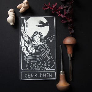 May include: A black and white illustration of Cerridwen, a figure from Welsh mythology, with a cauldron and flames. The image is on a black background with two small sculptures, a sprig of red leaves, and two wood carving tools.