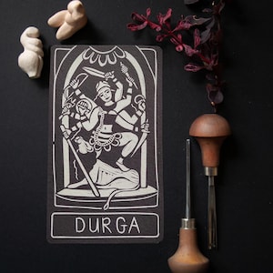 May include: A rectangular black and white linocut print featuring a multi-armed figure labeled DURGA at the bottom. The print is accompanied by two small light-colored abstract figurines and two wooden-handled carving tools with metal tips.