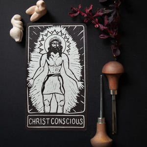 May include: A black and white linocut print featuring a central figure with a halo and the text CHRIST CONSCIOUS at the bottom. The print is accompanied by two wooden carving tools, two small light-colored carved figures, and a dark leafy branch.