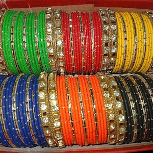 Könnte beinhalten: Eine Sammlung farbenfroher Armreifen in einer roten Box. Die Armreifen sind in verschiedenen Farben erhältlich, darunter Grün, Rot, Gelb, Blau, Orange und Schwarz. Jeder Armreif ist mit dekorativen goldfarbenen Akzenten und glitzernden Verzierungen versehen.