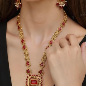 May include: A gold-toned necklace and earring set featuring red gemstone accents. The necklace has a large square pendant with a central red stone and smaller red teardrop-shaped stones. The earrings mirror the design with square red stones.