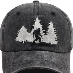 Könnte beinhalten: Eine schwarze, abgenutzte Baseballkappe mit einem gestickten Design eines Bigfoots, der durch einen Wald geht. Die Bäume sind weiß und der Bigfoot ist schwarz.