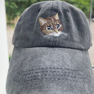 Könnte beinhalten: Eine schwarze Baseballkappe mit einem gestickten Katzenkopf auf der Vorderseite. Die Katze hat braunes Fell und grüne Augen.