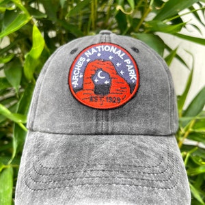 May include: A black, distressed, cotton baseball cap with an embroidered patch featuring the Arches National Park logo. The logo is a red circle with a white outline and the text "Arches National Park" in white. Inside the circle is a stylized image of Delicate Arch with a crescent moon and stars. The text "Est. 1929" is below the arch.