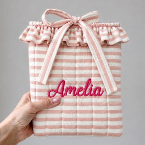 May include: A quilted pouch featuring a light pink and white horizontal striped pattern. The top edge has a ruffled trim and a matching fabric tie closure. The name Amelia is embroidered in a bright pink cursive script on the front center.