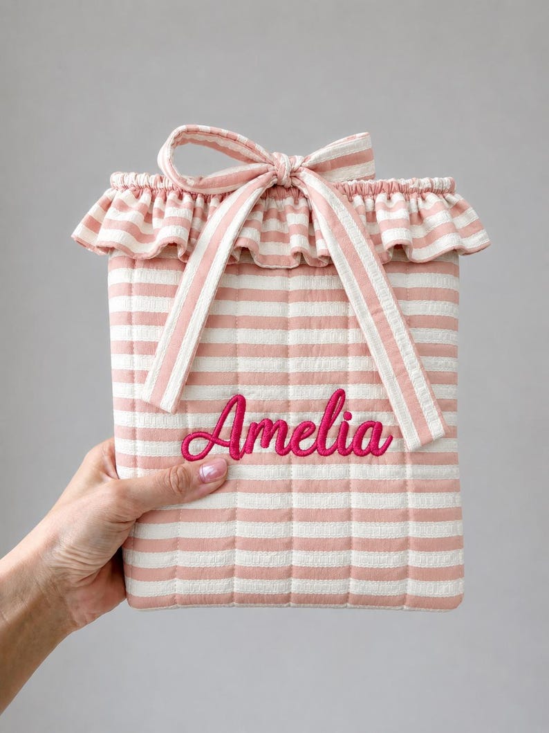 May include: A quilted pouch featuring a light pink and white horizontal striped pattern. The top edge has a ruffled trim and a matching fabric tie closure. The name Amelia is embroidered in a bright pink cursive script on the front center.