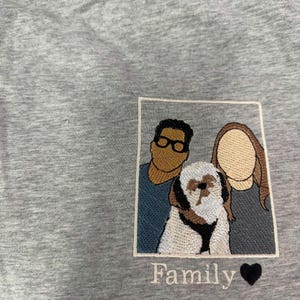 May include: A heather gray t-shirt featuring a rectangular embroidered patch. The patch depicts two faceless individuals and a small dog in the center. Below the patch, the word Family is embroidered in white script next to a black heart icon.