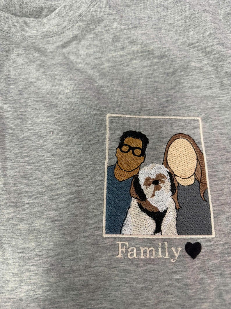 May include: A heather gray t-shirt featuring a rectangular embroidered patch. The patch depicts two faceless individuals and a small dog in the center. Below the patch, the word Family is embroidered in white script next to a black heart icon.