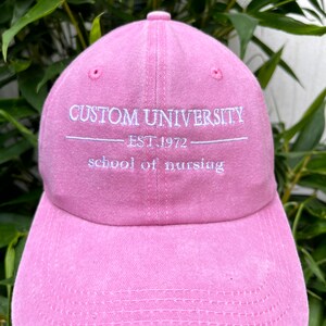 May include: A pink baseball cap with the text "CUSTOM UNIVERSITY EST. 1972 school of nursing" embroidered on the front.