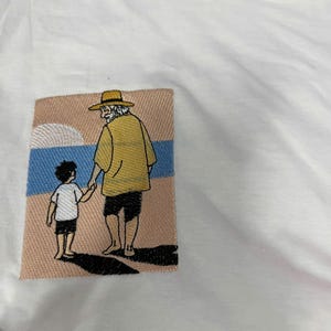 May include: A white t-shirt featuring a rectangular embroidered patch on the chest. The embroidery depicts an adult wearing a yellow shirt, black shorts, and a straw hat, holding hands with a child in a white shirt and black shorts on a beach.