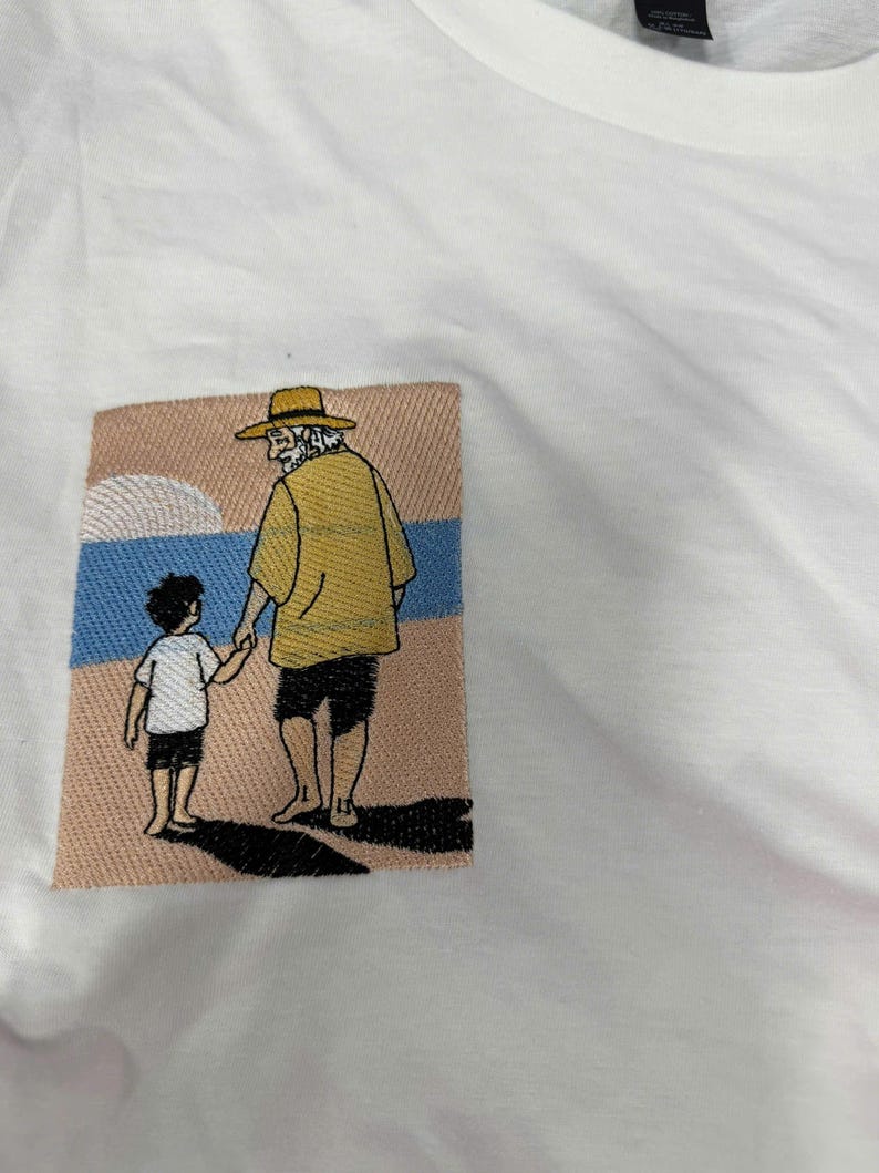 May include: A white t-shirt featuring a rectangular embroidered patch on the chest. The embroidery depicts an adult wearing a yellow shirt, black shorts, and a straw hat, holding hands with a child in a white shirt and black shorts on a beach.