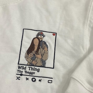May include: White sweatshirt with an embroidered image of a couple in a photo booth frame. The text "Wild Thing" and "The Troggs" is below the image, along with a music player icon.