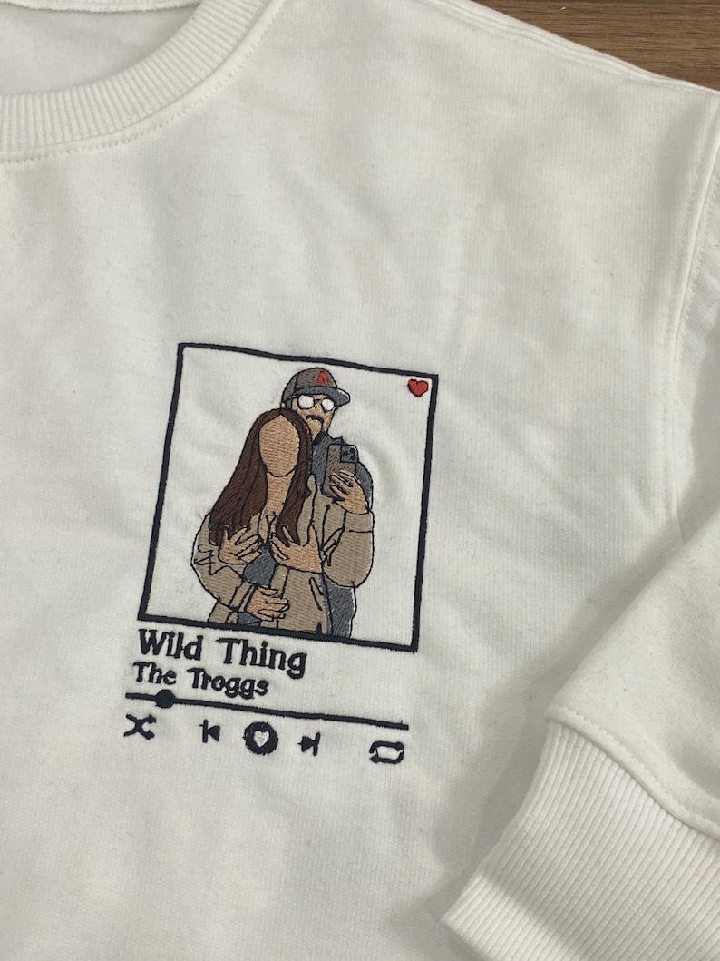 May include: White sweatshirt with an embroidered image of a couple in a photo booth frame. The text "Wild Thing" and "The Troggs" is below the image, along with a music player icon.