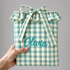 Könnte beinhalten: Eine gesteppte Tasche mit hellgrün-weißem Karomuster, einem gerüschten oberen Rand und einem Bindeband. Der Name Olivia ist in türkisfarbener Schreibschrift mittig auf der Vorderseite der rechteckigen Tasche eingestickt.