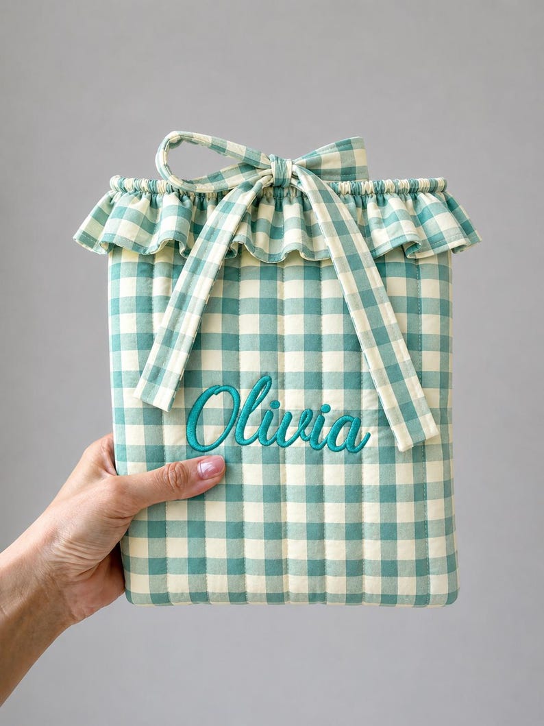 May include: A quilted pouch featuring a light green and white gingham pattern with a ruffled top edge and a fabric tie closure. The name Olivia is embroidered in teal script across the center of the front panel of the rectangular pouch.