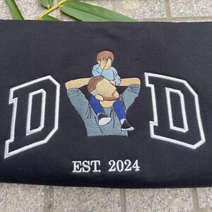K&ouml;nnte beinhalten: Schwarzer Sweatshirt mit wei&szlig;en gestickten Buchstaben, die "DAD" buchstabieren, mit einem kleinen gestickten Bild eines Vaters, der ein Kind auf seinen Schultern tr&auml;gt. Der Text "EST. 2024" ist unter dem Bild gestickt.