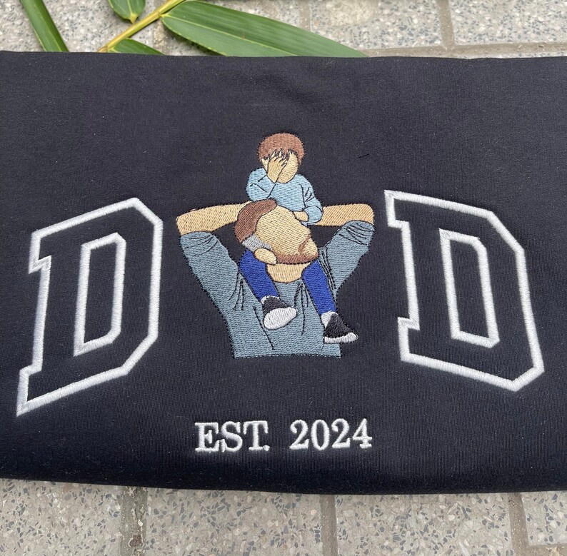 K&ouml;nnte beinhalten: Schwarzer Sweatshirt mit wei&szlig;en gestickten Buchstaben, die "DAD" buchstabieren, mit einem kleinen gestickten Bild eines Vaters, der ein Kind auf seinen Schultern tr&auml;gt. Der Text "EST. 2024" ist unter dem Bild gestickt.