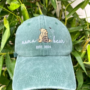 May include: Green baseball cap with an embroidered image of Winnie the Pooh holding a honey pot. The text "mama bear" and "EST 2024" is embroidered in white on the front of the cap.