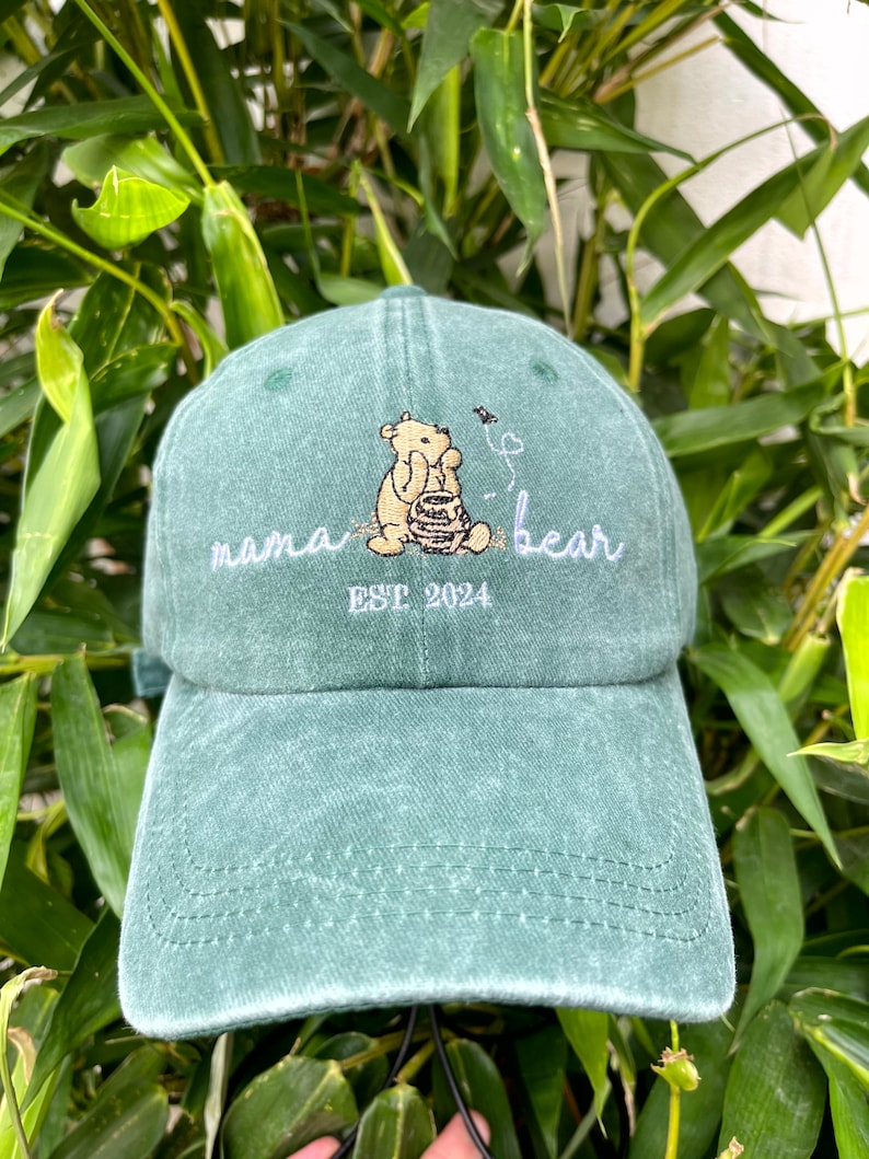 May include: Green baseball cap with an embroidered image of Winnie the Pooh holding a honey pot. The text "mama bear" and "EST 2024" is embroidered in white on the front of the cap.