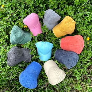May include: A collection of ten baseball caps in various colors including pink, gray, yellow, green, red, dark gray, light blue, blue, and beige. Each cap features a six-panel design with a curved brim and a button at the top of the crown.