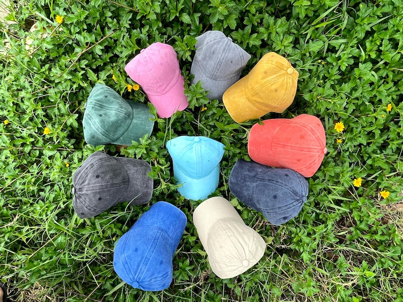 May include: A collection of ten baseball caps in various colors including pink, gray, yellow, green, red, dark gray, light blue, blue, and beige. Each cap features a six-panel design with a curved brim and a button at the top of the crown.