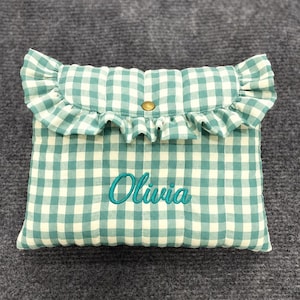 Könnte beinhalten: Eine gesteppte Tasche mit einem blaugrün-weißen Vichy-Muster, einer gerüschten Klappe und einem goldenen Druckknopfverschluss. Der Name Olivia ist in blaugrüner Schreibschrift auf der Vorderseite der rechteckigen Tasche eingestickt.