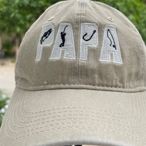 Könnte beinhalten: Eine beige Baseballkappe mit dem Wort "PAPA" in weiß gestickt und Angelmotiven in Schwarz.