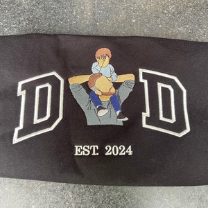 K&ouml;nnte beinhalten: Schwarzes besticktes T-Shirt mit einem wei&szlig;en Umriss des Wortes "DAD" und einem kleinen gestickten Bild eines Vaters, der ein Kind auf seinen Schultern tr&auml;gt. Der Text "EST. 2024" ist unter dem Bild gestickt.