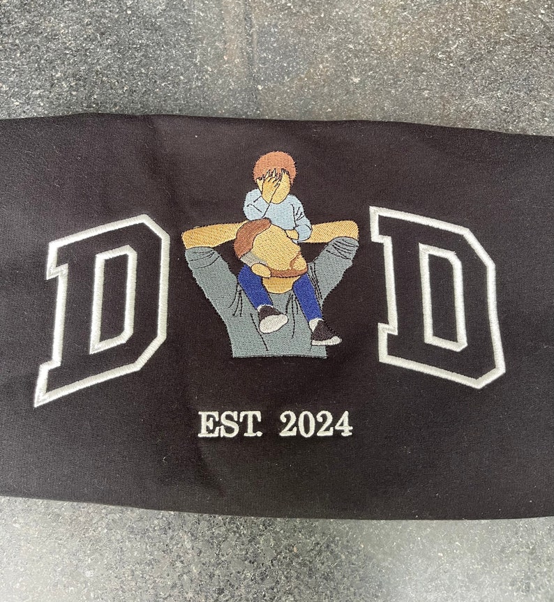 K&ouml;nnte beinhalten: Schwarzes besticktes T-Shirt mit einem wei&szlig;en Umriss des Wortes "DAD" und einem kleinen gestickten Bild eines Vaters, der ein Kind auf seinen Schultern tr&auml;gt. Der Text "EST. 2024" ist unter dem Bild gestickt.
