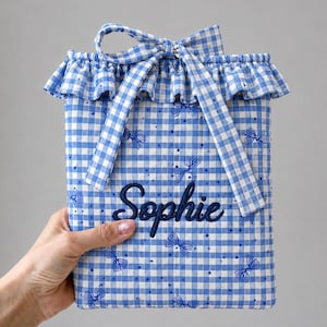 May include: A rectangular fabric pouch featuring a blue and white gingham pattern with small blue bow illustrations and dots. The pouch has a ruffled top edge, a tied fabric bow closure, and the name Sophie embroidered in dark blue script.