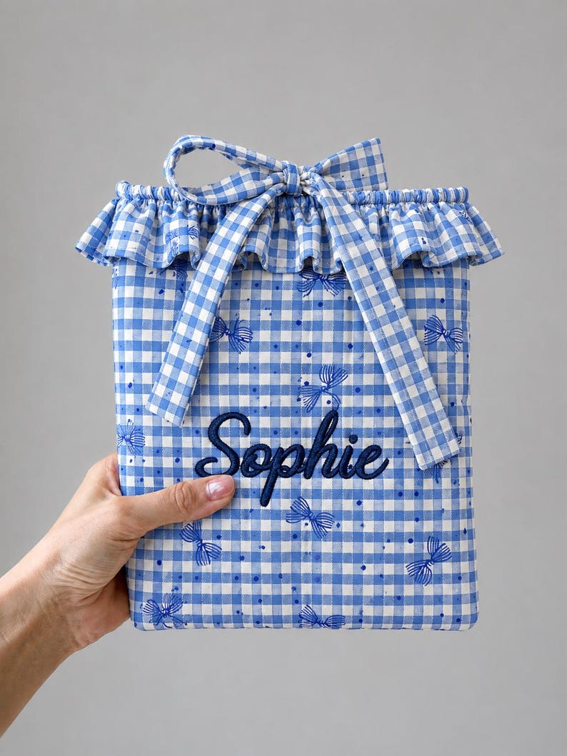 May include: A rectangular fabric pouch featuring a blue and white gingham pattern with small blue bow illustrations and dots. The pouch has a ruffled top edge, a tied fabric bow closure, and the name Sophie embroidered in dark blue script.