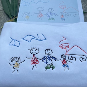 Könnte beinhalten: Ein weißer Pullover mit einer bunten gestickten Zeichnung einer Familie mit einem Haus und einem Auto. Die Zeichnung ähnelt einer Kinderzeichnung.