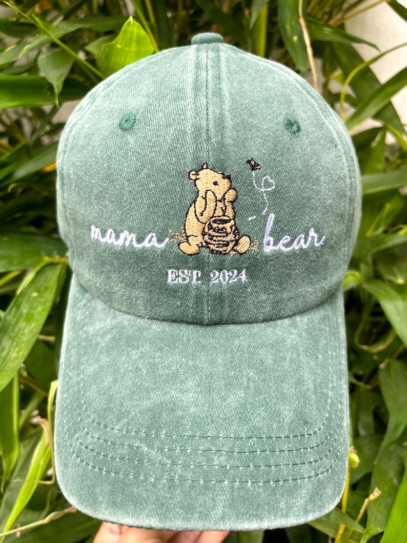 May include: Green baseball cap with an embroidered image of Winnie the Pooh holding a honey pot. The text "mama bear" and "EST 2024" is also embroidered on the cap.