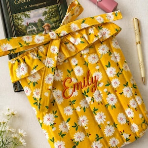 May include: A quilted yellow fabric sleeve featuring a white daisy pattern with green leaves, a ruffled top edge, and a tie closure. The name Emily is embroidered in orange script on the front. The sleeve is positioned next to a book, a pen, and a lipstick.