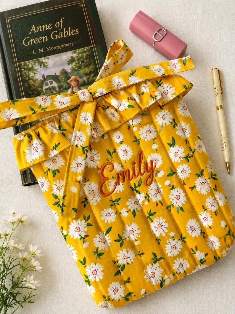 May include: A quilted yellow fabric sleeve featuring a white daisy pattern with green leaves, a ruffled top edge, and a tie closure. The name Emily is embroidered in orange script on the front. The sleeve is positioned next to a book, a pen, and a lipstick.