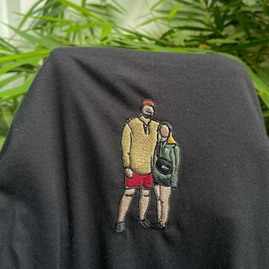 May include: Embroidered design on a black t-shirt featuring a couple standing side-by-side. The man is wearing a yellow shirt, red shorts, and a brown hat. The woman is wearing a green shirt and black shorts.