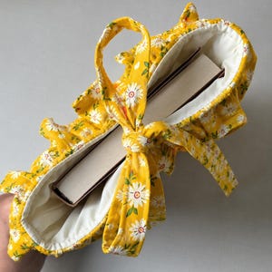 May include: A bright yellow fabric book sleeve featuring a white daisy floral pattern with green leaves. The sleeve has a ruffled edge, a white interior lining, and a matching fabric tie closure that is knotted at the top to secure a thick book inside.