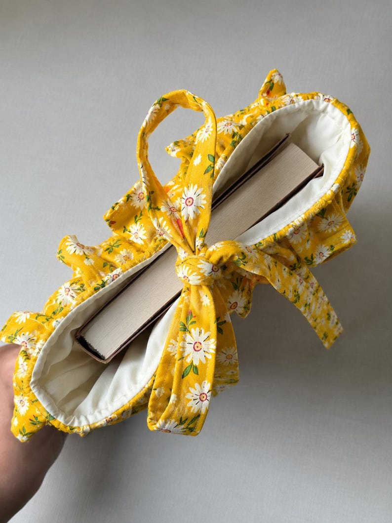 May include: A bright yellow fabric book sleeve featuring a white daisy floral pattern with green leaves. The sleeve has a ruffled edge, a white interior lining, and a matching fabric tie closure that is knotted at the top to secure a thick book inside.