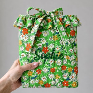 May include: A quilted fabric pouch featuring a vibrant green background with a repeating pattern of white, yellow, and orange daisy flowers. The pouch has a ruffled top edge, a tie closure, and the name Sophie embroidered in dark green script.