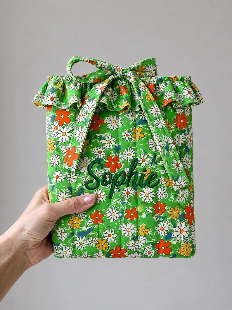 May include: A quilted fabric pouch featuring a vibrant green background with a repeating pattern of white, yellow, and orange daisy flowers. The pouch has a ruffled top edge, a tie closure, and the name Sophie embroidered in dark green script.