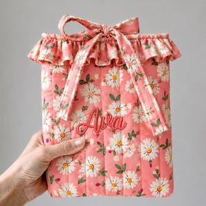May include: A quilted fabric pouch in a coral pink hue featuring a white daisy floral pattern with green leaves. The pouch has a ruffled top edge, a matching fabric tie closure, and the name Ava embroidered in dark pink script on the front.