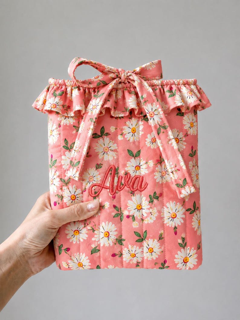 May include: A quilted fabric pouch in a coral pink hue featuring a white daisy floral pattern with green leaves. The pouch has a ruffled top edge, a matching fabric tie closure, and the name Ava embroidered in dark pink script on the front.