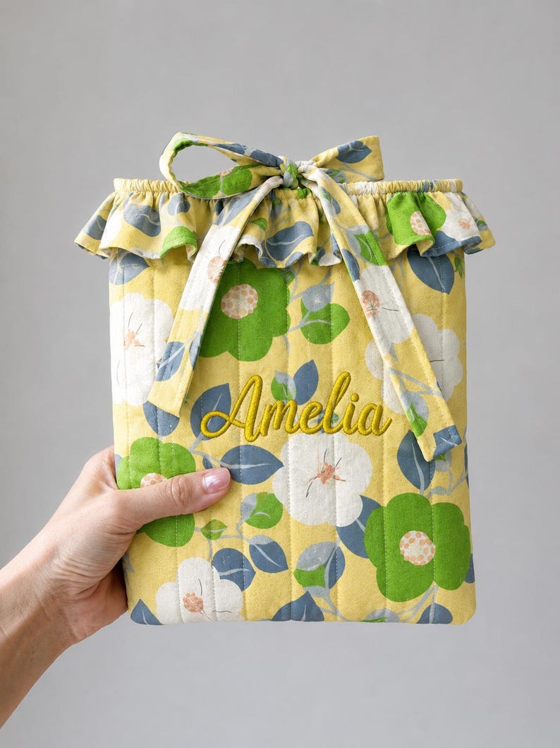 May include: A quilted fabric pouch in yellow featuring a floral pattern of green and white flowers with blue leaves. The pouch has a ruffled top edge, a tie closure, and the name Amelia embroidered in yellow thread on the front surface.